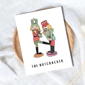 Könnte beinhalten: Eine weiße Karte mit einer Illustration von zwei Nussknackern. Einer, in Rot und Grün, hält eine Trommel. Der andere, in Grün und Gold, wird spielerisch getreten. Der Text "THE NUTCRACKER" ist darunter gedruckt.