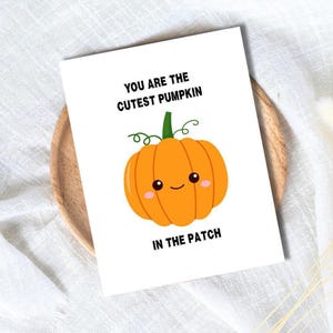Pode incluir: Um cartão branco com o texto "YOU ARE THE CUTEST PUMPKIN IN THE PATCH" e uma ilustração de desenho animado de uma abóbora laranja com um rosto sorridente. O cartão está em um prato de madeira.