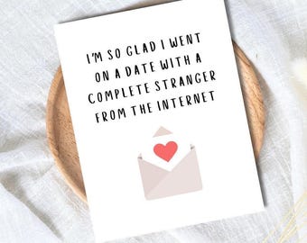 May include: A white greeting card with the text "I'M SO GLAD I WENT ON A DATE WITH A COMPLETE STRANGER FROM THE INTERNET." An illustration of an envelope with a red heart is below the text. The card is on a wooden plate.
