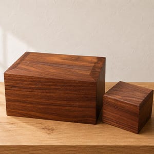 May include: Two rectangular wooden boxes made of dark brown walnut with a visible natural wood grain pattern. The larger box is positioned on the left, while a smaller, square-shaped box sits to its right, both featuring smooth, flat surfaces.