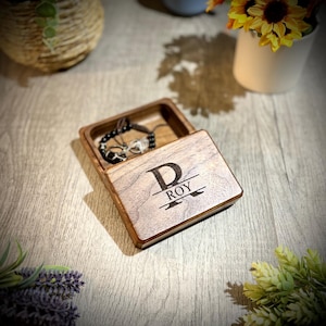 May include: A wooden box with a hinged lid. The lid is engraved with the letter "D" and the name "ROY". The box is open and a black beaded bracelet is inside.