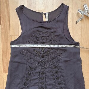 May include: A dark gray sleeveless dress with intricate beaded embellishments. The dress features a keyhole neckline and a flowing skirt. A measuring tape shows the dress is approximately 16 inches wide. The dress is on a wooden surface.