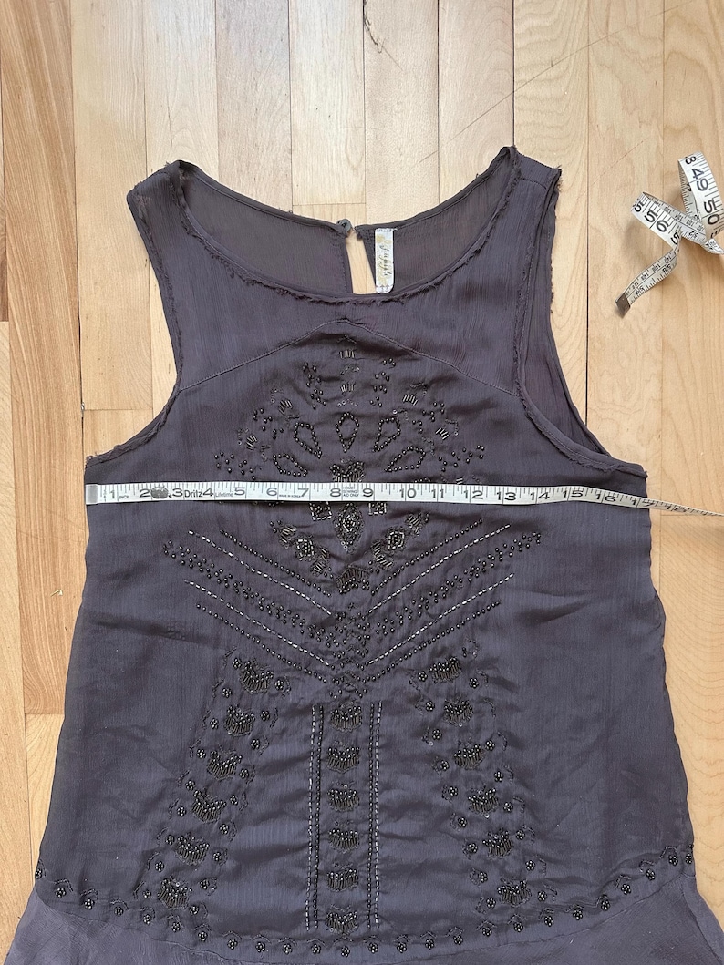 May include: A dark gray sleeveless dress with intricate beaded embellishments. The dress features a keyhole neckline and a flowing skirt. A measuring tape shows the dress is approximately 16 inches wide. The dress is on a wooden surface.