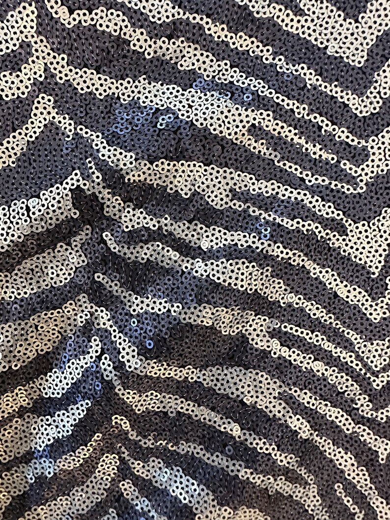 May include: Close-up of a fabric with a sequined zebra stripe pattern. The sequins are in shades of silver and navy blue, creating a shimmering effect. The design features a repeating chevron pattern.