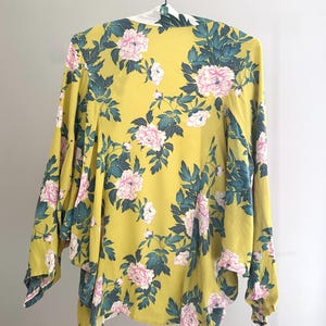 May include: A yellow floral kimono with pink peony flowers and dark green leaves. The kimono is hanging on a white hanger against a white background. The sleeves are long and flowing, and the fabric appears to be lightweight.