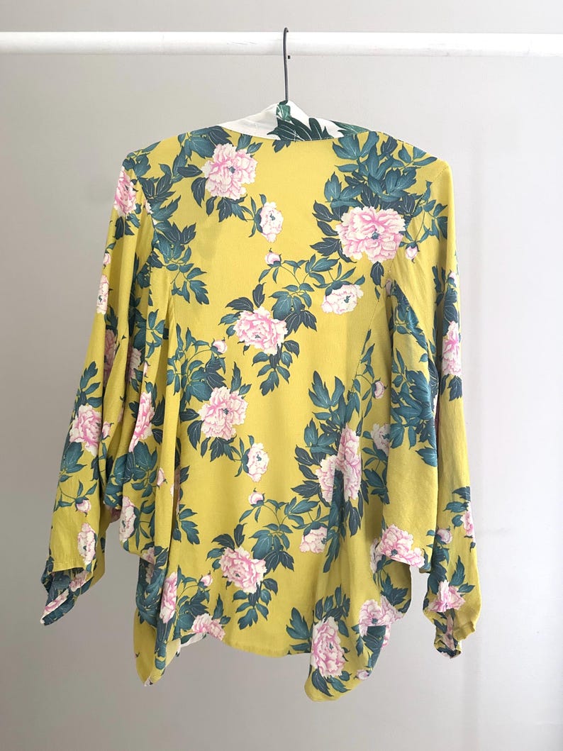 May include: A yellow floral kimono with pink peony flowers and dark green leaves. The kimono is hanging on a white hanger against a white background. The sleeves are long and flowing, and the fabric appears to be lightweight.