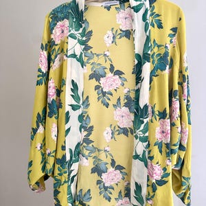May include: A yellow kimono jacket featuring a floral design of pink peonies and green foliage. The jacket has long sleeves and a white border. The garment is suspended from a wooden hanger.