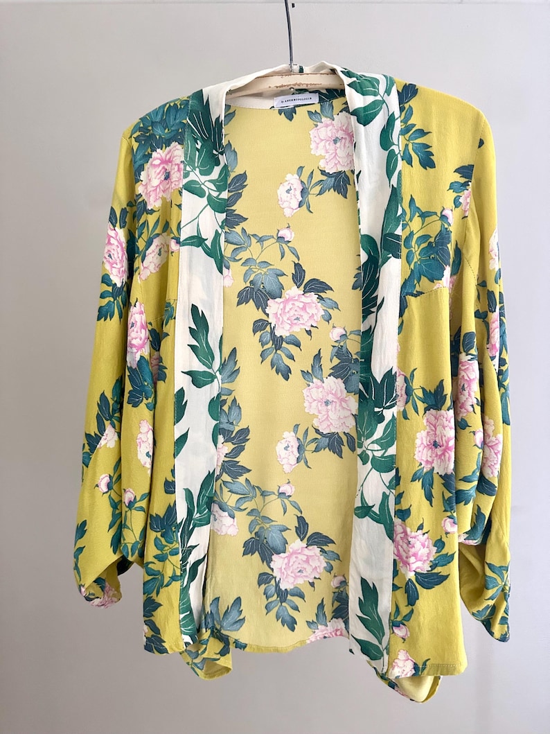 May include: A yellow kimono jacket featuring a floral design of pink peonies and green foliage. The jacket has long sleeves and a white border. The garment is suspended from a wooden hanger.
