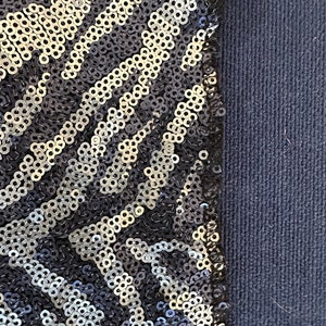 May include: Close-up of a section of fabric with a sequined pattern. The sequins are silver and black, arranged in a diagonal zebra stripe design. The fabric on the right is a solid navy blue.