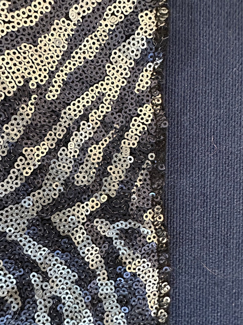 May include: Close-up of a section of fabric with a sequined pattern. The sequins are silver and black, arranged in a diagonal zebra stripe design. The fabric on the right is a solid navy blue.