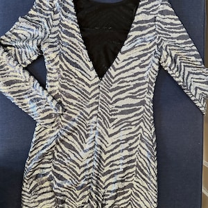 May include: A silver and black sequined dress with a zebra stripe pattern. The dress has long sleeves and a deep V-neck in the front and a low back. The back of the dress is black mesh. The dress is laying flat on a dark blue surface.