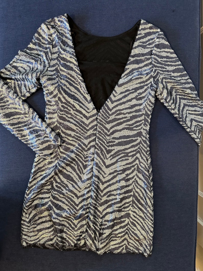 May include: A silver and black sequined dress with a zebra stripe pattern. The dress has long sleeves and a deep V-neck in the front and a low back. The back of the dress is black mesh. The dress is laying flat on a dark blue surface.