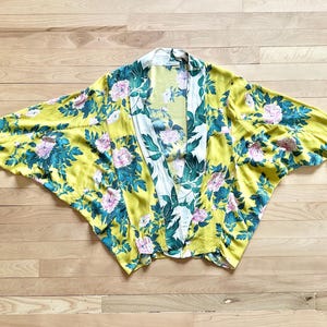 May include: Yellow floral kimono with a white and green floral pattern on the lapel. The kimono features a vibrant design of pink flowers and teal leaves. The garment is made of a lightweight fabric and is spread out on a wooden surface.