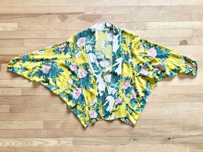May include: Yellow floral kimono with a white and green floral pattern on the lapel. The kimono features a vibrant design of pink flowers and teal leaves. The garment is made of a lightweight fabric and is spread out on a wooden surface.