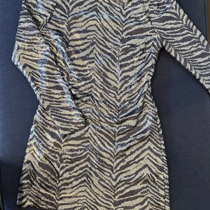 May include: A long-sleeved mini dress with a zebra stripe pattern. The dress is covered in silver sequins over a dark blue fabric. The dress has a scoop back and is laying on a dark blue surface.