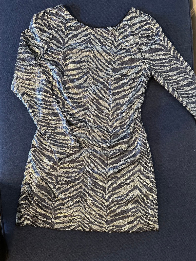 May include: A long-sleeved mini dress with a zebra stripe pattern. The dress is covered in silver sequins over a dark blue fabric. The dress has a scoop back and is laying on a dark blue surface.