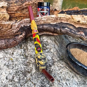 Könnte beinhalten: Eine dunkelbraune Holzpfeife mit einem perlenbesetzten Abschnitt in Gelb, Rot und Schwarz, einem kleinen violetten Akzent und einer geformten beigen Tierkopfperle. Ein kleines Glas mit der Aufschrift CEREMONIAL und eine dunkle Schale mit braunem Pulver.