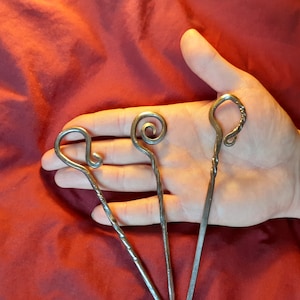 Könnte beinhalten: Drei Haarnadeln aus Metall mit verschiedenen dekorativen Enden. Die Nadeln werden in einer Hand vor einem roten Hintergrund gehalten.