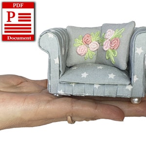 May include: A miniature blue velvet sofa with white stars and a floral embroidered cushion. The sofa is sitting on a hand.