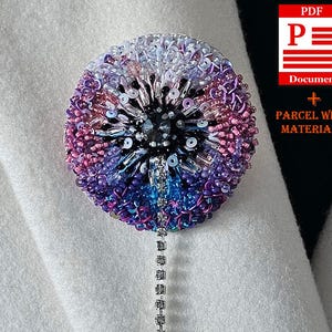 Könnte beinhalten: Ein runder, mit Perlen besetzter Broschenanhänger in Lila, Blau und Weiß. Die Brosche hat einen dunklen Mittelstein und eine herabhängende Kette aus kleinen, klaren Steinen. Die Brosche ist an einem weißen Stoff befestigt.