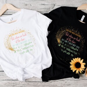 May include: Two t-shirts, one white and one black, with the same text design. The design features a gold glitter crescent and text that reads "WE ARE MERE MORTALS But when we look at the stars..."