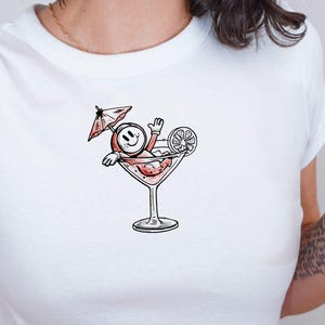 May include: White, short-sleeved crop top with a cartoon illustration of a smiley face relaxing in a martini glass. The design includes a cocktail umbrella and a lemon slice. The top is made of a soft material.