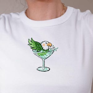 May include: White crop top with a quirky design of an eagle in a martini glass. The eagle has green wings and a white head, and the glass is filled with green liquid. The top is a casual, short-sleeved style, perfect for a relaxed look.
