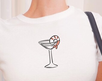 May include: A white short-sleeved crew neck t-shirt featuring a minimalist black line drawing of a martini glass with a pink and black shrimp garnish centered on the chest. The shirt has a fitted silhouette and a simple hemline.