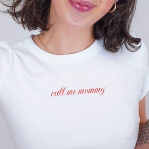 May include: White crop top with the phrase "call me mommy" in red script. The shirt is paired with light wash blue jeans. A martini glass with an olive is held in the hand.