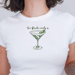 May include: White crop top with short sleeves featuring a green martini glass with olives and the text "the bride needs a".