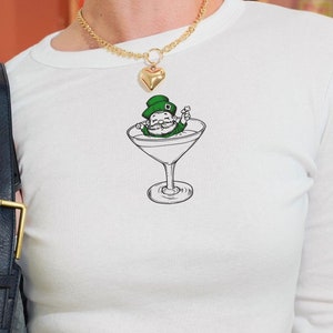 May include: White long-sleeve shirt with a green leprechaun in a martini glass graphic. The shirt has a ribbed texture and a crew neckline.