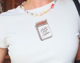 May include: A white short-sleeved t-shirt featuring a graphic of an open matchbox with the text Match Made in Heaven. The shirt is paired with a multicolored beaded necklace and a black leather shoulder bag over gray trousers.