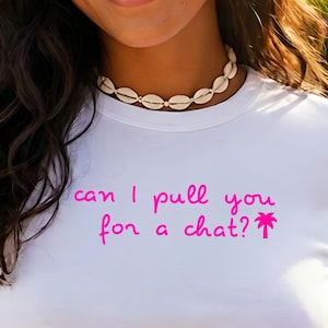 May include: A white short-sleeved crew neck t-shirt featuring the pink text can I pull you for a chat? accompanied by a small pink palm tree icon. The shirt is made of a plain fabric and has a fitted silhouette with a standard hemline.