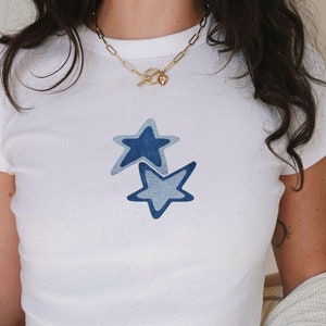 May include: White cropped t-shirt featuring a blue and light blue star design. The shirt is worn with a white cardigan and grey sweatpants.