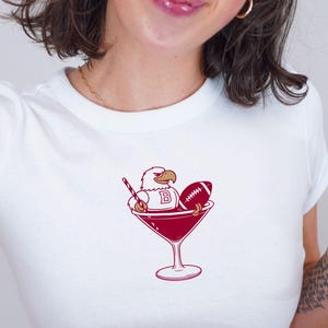 May include: White crop top featuring a maroon graphic of a martini glass with an eagle, football, and a stir stick. The eagle has a letter "B". The shirt is worn with blue jeans.