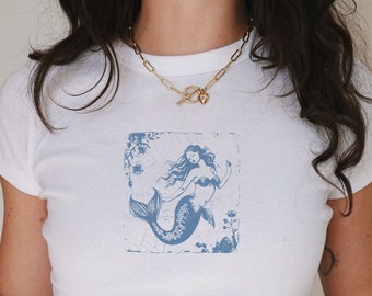May include: A white short-sleeved t-shirt featuring a blue graphic print of a mermaid centered on the chest. The mermaid design is enclosed in a square frame with floral accents. The shirt is paired with a gold-toned chain link necklace.