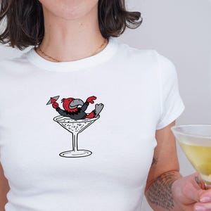 May include: White crop top featuring a cartoon bird in a martini glass. The bird is red, black, and grey. The person is wearing blue jeans and holding a martini glass with an olive.