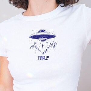 May include: White crop top with a graphic of a UFO, mountains, and a person with the text "FINALLY" printed on the front.