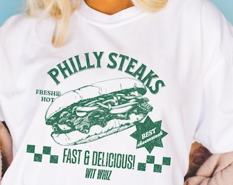 May include: White t-shirt with a green graphic of a Philly cheesesteak sandwich. The graphic says "Philly Steaks", "Fresh & Hot", "Best Cheesesteak", "Fast & Delicious!" and "Wit Whiz".