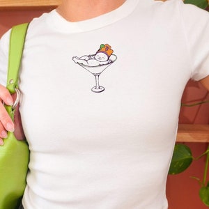 May include: White t-shirt with a graphic of a baby wearing a crown and lying in a martini glass.