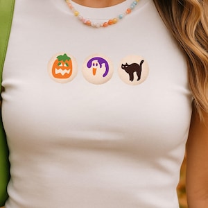May include: A white short-sleeved t-shirt featuring three circular graphics across the chest: an orange jack-o'-lantern, a purple ghost, and a black cat. The shirt is paired with a bright green shoulder bag and a colorful beaded necklace.