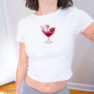May include: White, short-sleeved t-shirt with a maroon graphic of an eagle, football, and cocktail glass. The eagle holds a pencil, and the football has a letter "B". The graphic is centered on the chest.