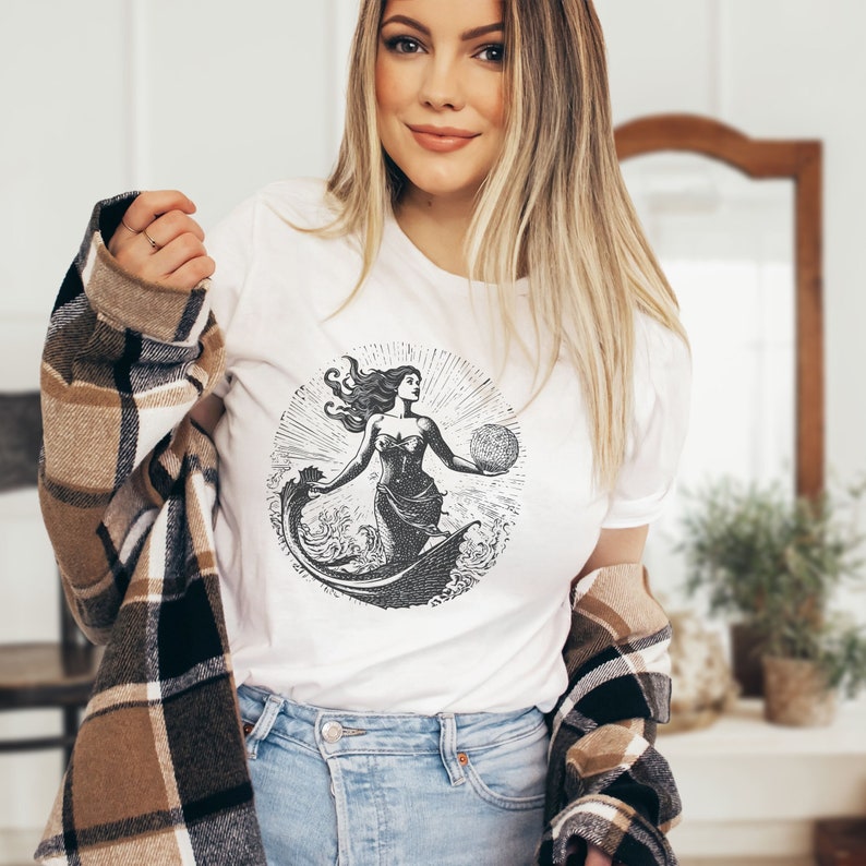 May include: White t-shirt with a black and white graphic print of a mermaid holding a globe. The graphic is in a circle with a sunburst design around it.