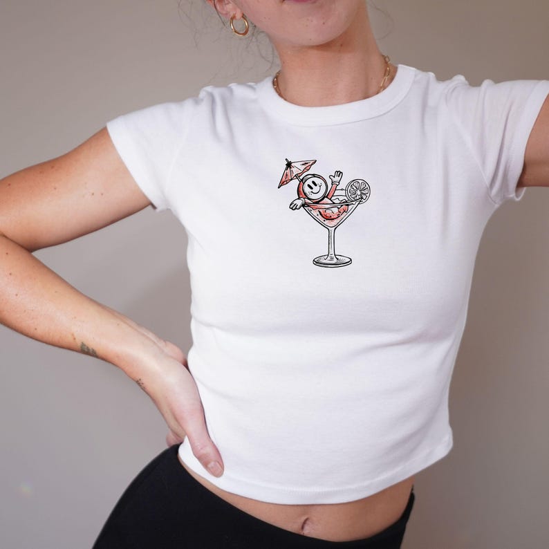 May include: White, short-sleeved crop top with a cartoon graphic of a smiley face relaxing in a cocktail glass, complete with a miniature umbrella and a lemon slice. The graphic is in red, black, and white.