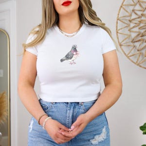 Puede incluir: Camiseta blanca recortada con un gr&aacute;fico de una paloma con un lazo rosa.