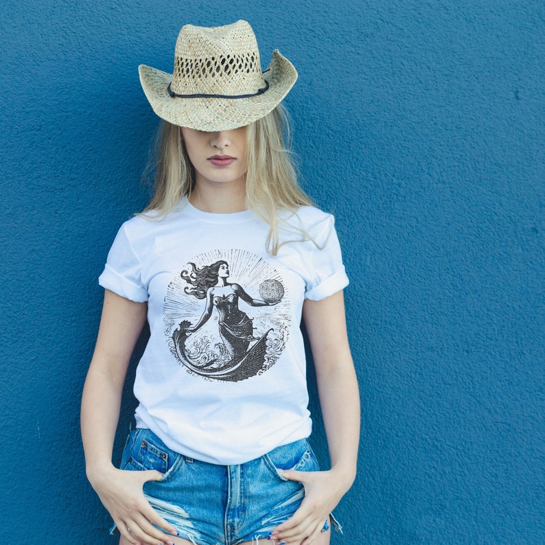 May include: A white t-shirt with a black and white illustration of a mermaid holding a globe. The mermaid is surrounded by waves. The t-shirt is being worn by a person with long blonde hair and a straw cowboy hat.