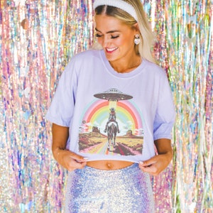 Op de afbeelding: Een lavendelkleurig gecropped t-shirt met een afbeelding van een cowboy die een paard rijdt onder een regenboog met een UFO erboven. De afbeelding is in retro-stijl.