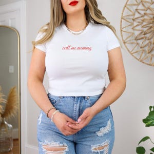 May include: A white, short-sleeved t-shirt with the red script text "call me mommy". The top is worn with blue denim jeans. A gold chain necklace and bracelets are also visible. The outfit has a casual, relaxed style.