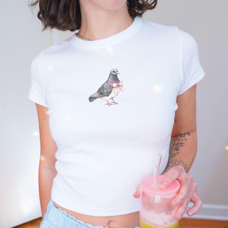 Puede incluir: Una camiseta blanca recortada con un gr&aacute;fico de una paloma con un lazo rosa. La paloma est&aacute; mirando hacia la izquierda y tiene un esquema de color gris y blanco.