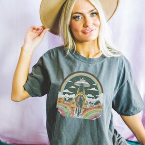 May include: A dark gray t-shirt with a graphic print of a woman riding a horse with a rainbow and a UFO in the background. The graphic is in a retro style with bright colors.
